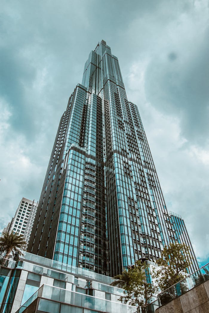 hero-img-01 A towering, modern skyscraper with a glass facade under a dramatic cloudy sky, showcasing urban architecture.